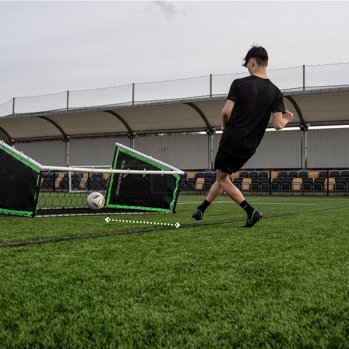 TEKKERS GS Trainer laag opgesteld op een kunstgrasveld terwijl een bal binnenrolt, met een speler die passt en een stippelpijl voor de terugkaatsing.