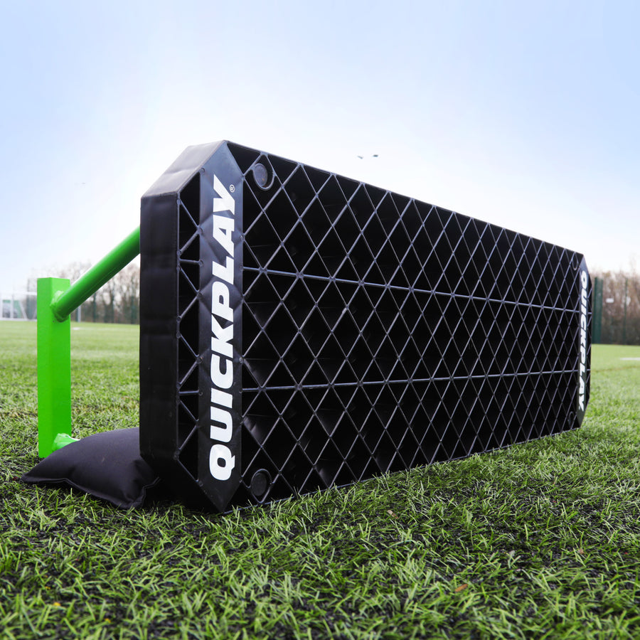REPLAY Station XL reboundbord op kunstgras, laag zijaanzicht met de zwarte roostervoorkant, het groene frame en de zwarte tas.