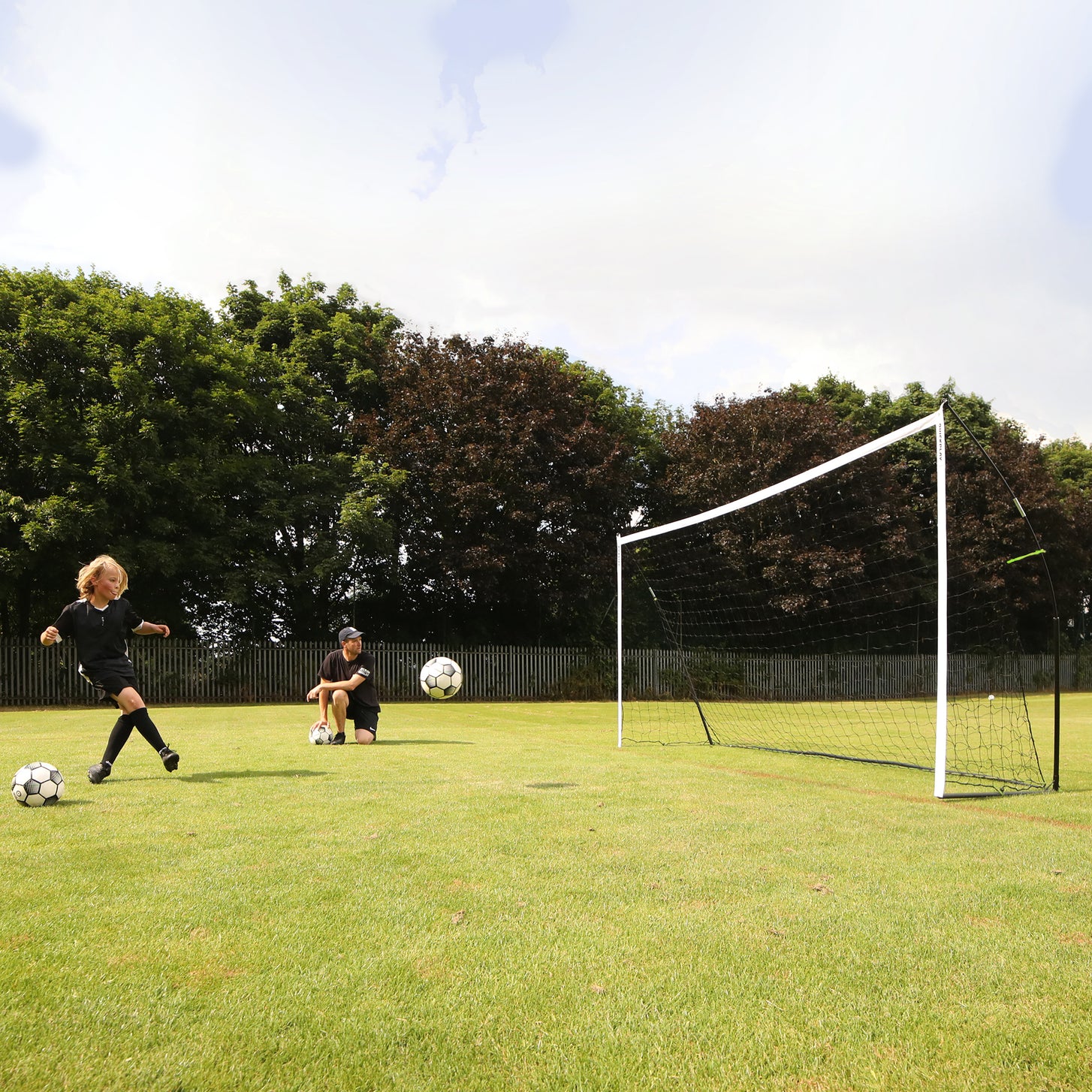 Kind schiet op het KICKSTER doel 500 x 200 cm op het gras terwijl een geknielde volwassene toekijkt, met een bal in de lucht.