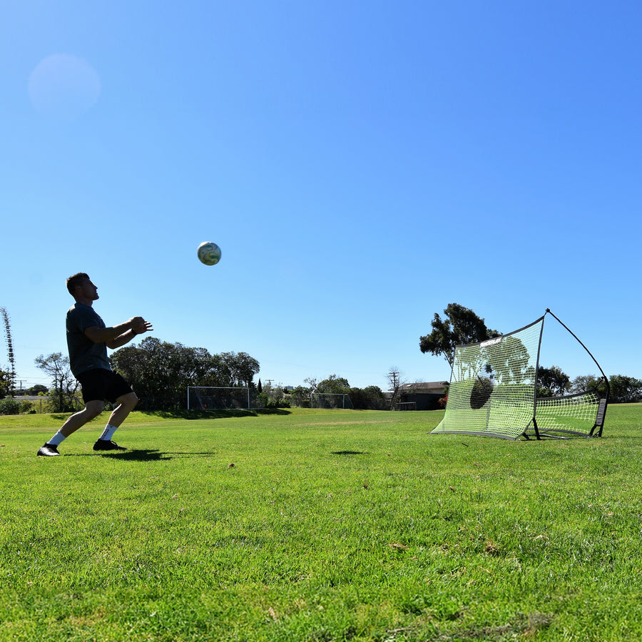 Breed beeld van een speler die zich klaarmaakt om een bal in de lucht aan te nemen, met de TEKKERS Trainer rebounder achter hem op het gras.